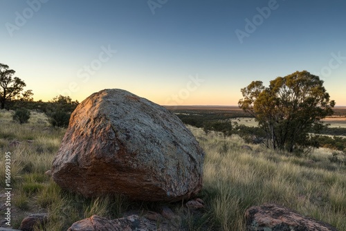 The venom rock in the outback of Australia, an isolated red boulder at dusk with tree silhouettes in the background. The sky is clear and blue. The photo was taken from behind the boulders  photo