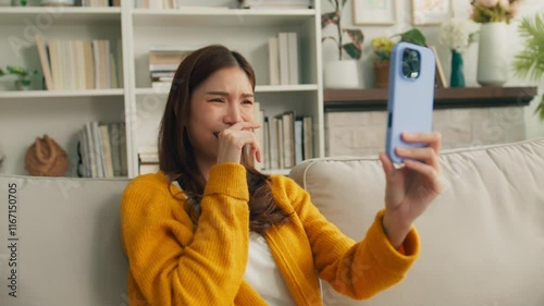 Happy young Asian woman looking at mobile phone connects online conversations with friends through a video chat app on smartphone enjoying online communication sitting on sofa in a cozy room at home.