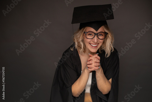 A joyful graduate in a black cap and gown, wearing glasses, leans forward with clasped hands and a beaming smile, expressing excitement and accomplishment against a neutral background. photo