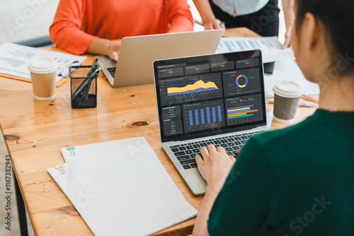 team collaborates in office, analyzing data on laptop with charts and graphs, ed by documents, coffee cups, and stationery on wooden desk photo