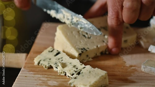 Close-up of hands slicing blue cheese on a wooden board. Preparing a cheese plate for dinner or a party. Soft lighting with bokeh adds a touch of elegance.
