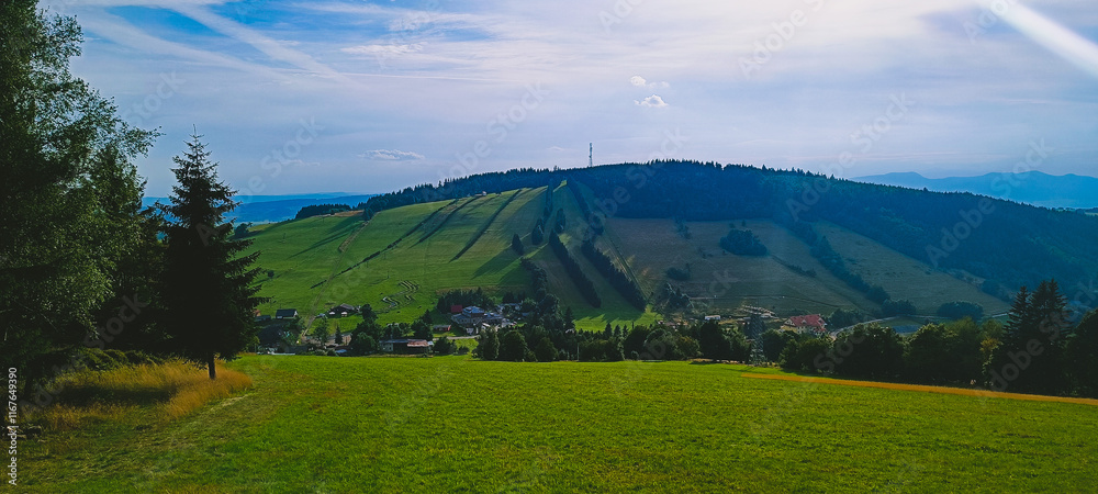 Naklejka premium Zachwycający krajobraz górski z zielonymi łąkami, lasami i wioską u podnóża wzgórza. Na szczycie wzgórza widoczny jest maszt telekomunikacyjny, co dodaje nowoczesnego akcentu naturalnemu pejzażowi