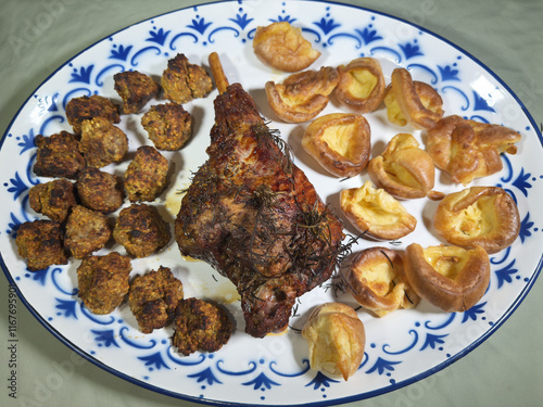 A whole leg of hot freshly roasted lamb on a large serving plate, covered in rosemary herb, and surrounded by Yorkshire puddings and stuffing balls. photo