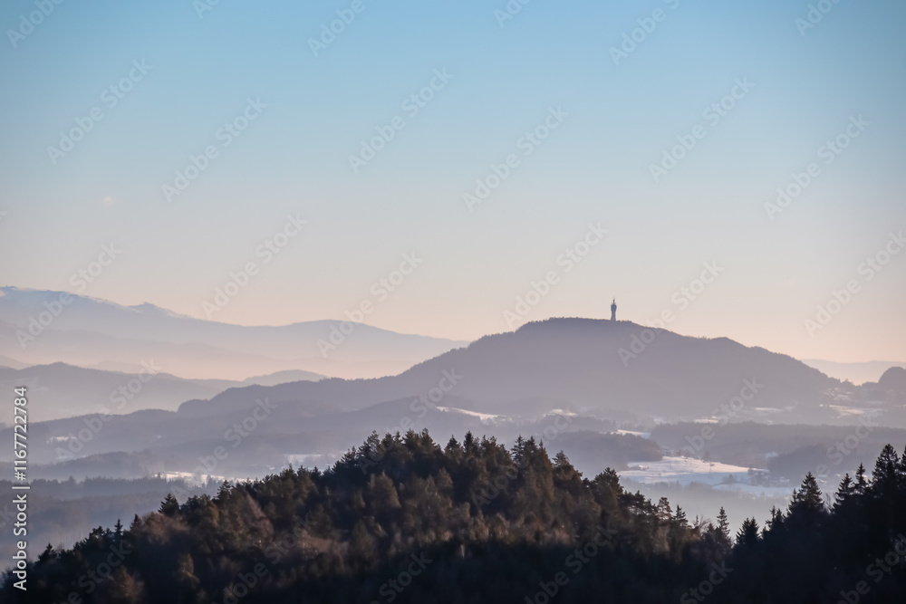 Obraz premium Panoramic view of Pyramidenkogel in Rosental Valley in winter seen from mount Petelin in Faak, Carinthia, Austria. Alpine landscape in soft light. Austrian Alps shrouded in mist. Serenity and calmness