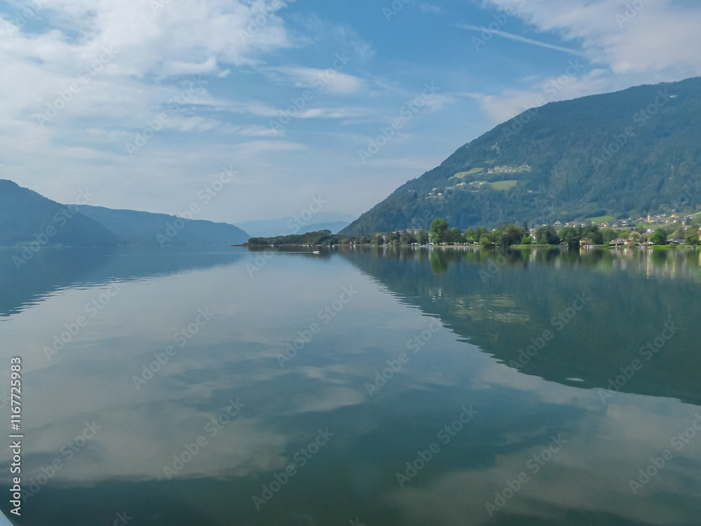 Breathtaking view of Lake Ossiach with water reflections of forest covered hills of Ossiacher Tauern in Carinthia, Austria, Europe. Clear blue sky in summer. Serene and idyllic scene in Austrian Alps Breathtaking view of Lake Ossiach with water reflections of forest covered hills of Ossiacher Tauern in Carinthia, Austria, Europe. Clear blue sky in summer. Serene and idyllic scene in Austrian Alps