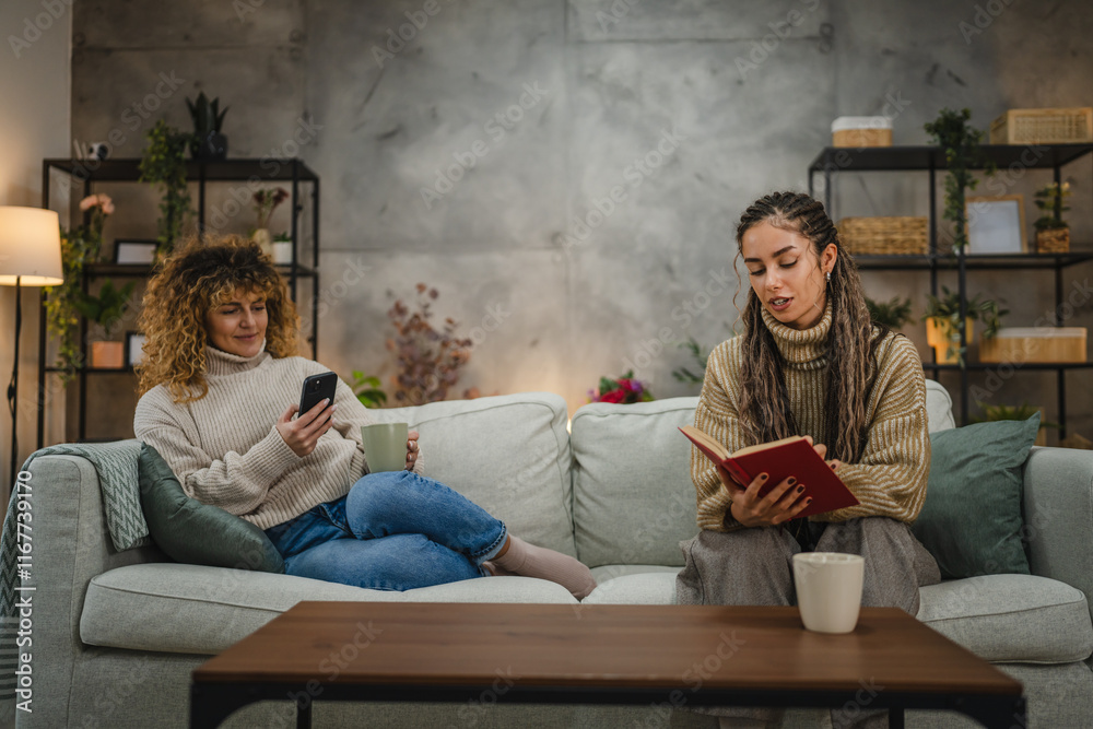 Obraz premium female friends enjoy on sofa one read a book while other use cellphone
