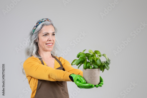 A mature woman wearing a vibrant outfit and gardening gloves holds a potted plant with pride. Her joyful demeanor reflects her hobby of gardening, showcasing her love for plants. photo