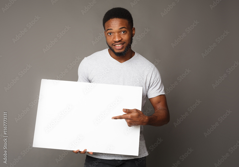 Picture of young smiling african-american man holding white blank board and pointing on it, on grey background, copy space Picture of young smiling african-american man holding white blank board and pointing on it, on grey background, copy space