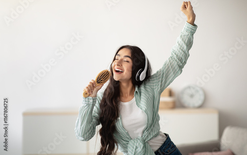 Fotografie Happy Indian lady with headphones dancing to music and singing song, using hairbrush as mic at home