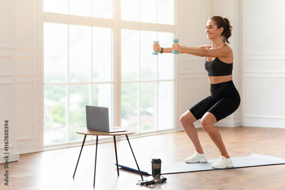 Obraz premium Positive fit lady exercising and squatting with two dumbbells in front of pc on coffee table at home, smiling fitness blogger recording video for her followers having personal virtual training