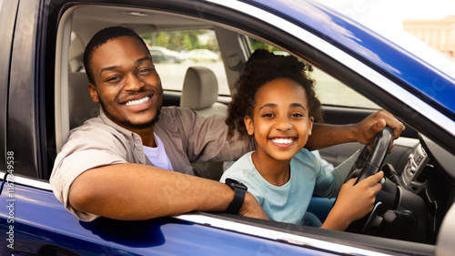 Happy Black Daddy Teaching Preteen Daughter To Drive Smiling To Camera, Sitting In Driver's Seat With Father Together. Child Learning To Drive Auto Having Fun Traveling By Automobile