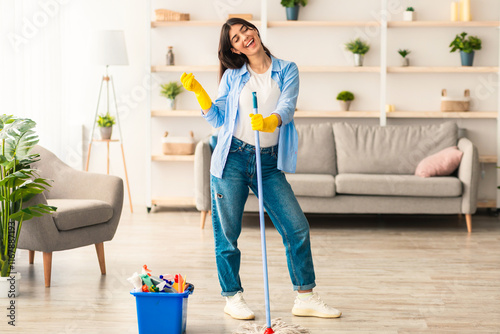 Happy young woman cleaning her flat, singing and listening to music. Excited maid dancing and holding mop, having fun, free space. Millennial housewife in protective gloves enjoying domestic chores