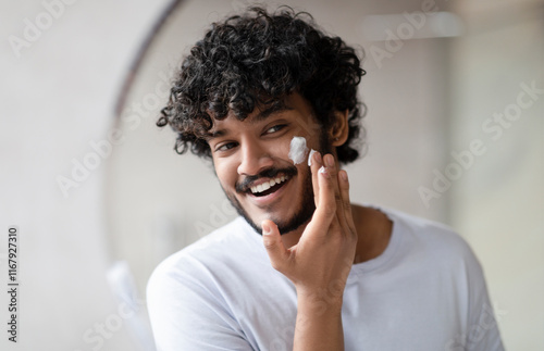 Facial skincare. Indian man applying moisturizing cream on face, looking at his reflection in bathroom mirror at home, free space. Male beauty and skin care concept