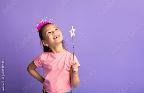 Cheerful asian girl wearing princess crown on head, holding magic wand, looking up, pastel purple studio wall, copy space photo