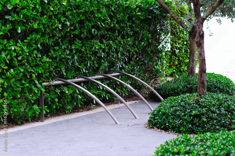 Naklejka premium Bike rack parking lot with curved metal bars near green tropical and paved walkway. Urban city life.
