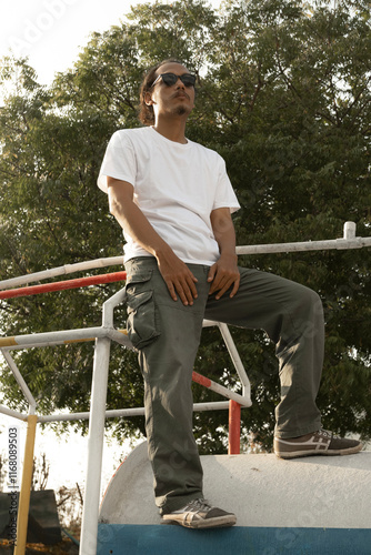 An Asian male model wearing a plain white t-shirt and black sunglasses is posing in a commercial park during the daytime. The male model's white t-shirt is being used as a mockup object for showcasing