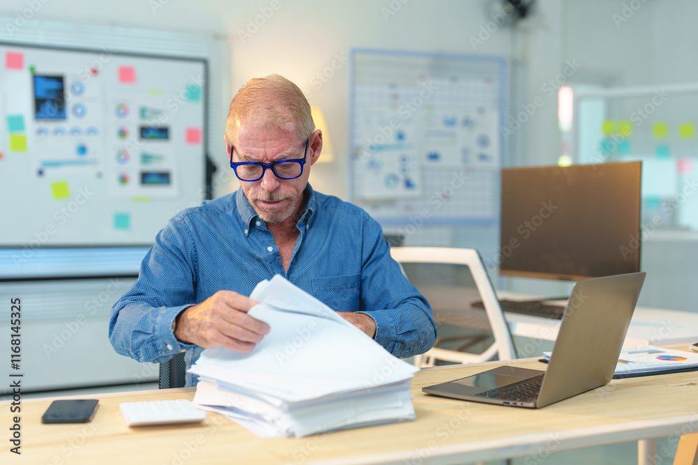 Focused mature manager examining documents, working with laptop and financial reports in modern office, analyzing data and preparing presentation Focused mature manager examining documents, working with laptop and financial reports in modern office, analyzing data and preparing presentation