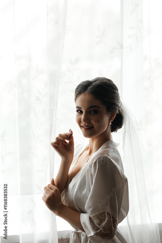 the bride stands by the window with translucent curtains with a floral pattern. She is dressed in a white robe with lace inserts and holds a ring in her hands. the bride stands by the window with translucent curtains with a floral pattern. She is dressed in a white robe with lace inserts and holds a ring in her hands.