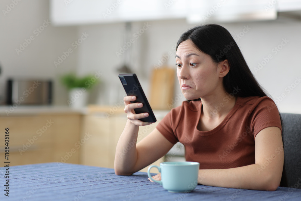 Perplexed asian woman checking phone in the kitchen Perplexed asian woman checking phone in the kitchen