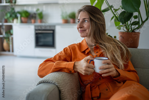 Cheerful woman enjoys coffee or tea on couch, taking break in cozy home living room with houseplants. Female couch potato, slow living, relaxed weekend, hygge alone, carefree leisure, home relaxation photo