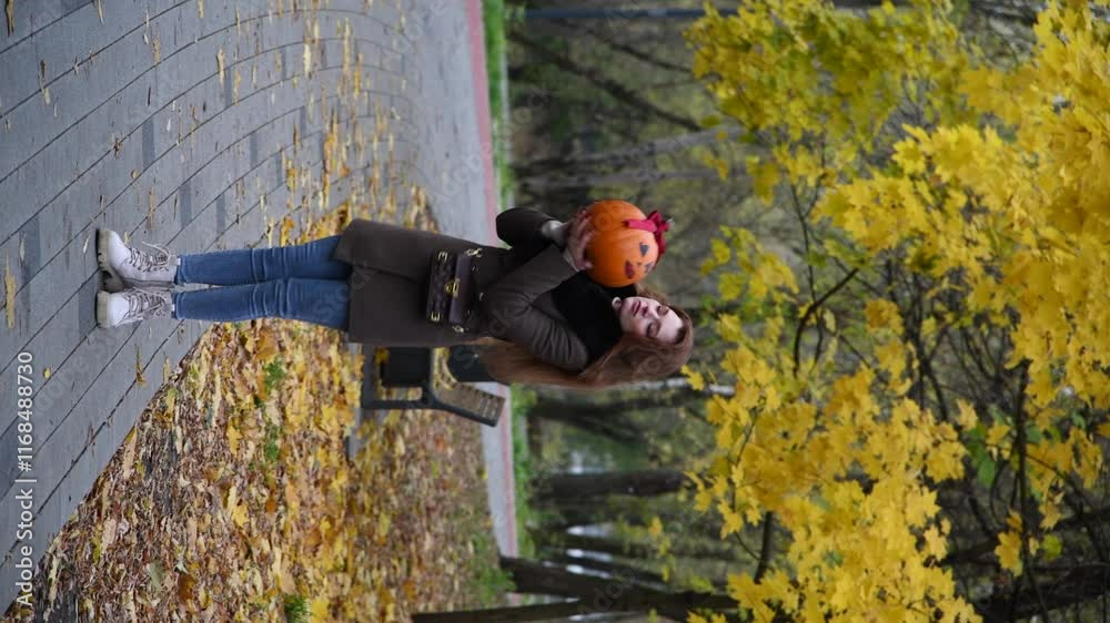 Portrait of beautiful brunette woman in brown jacket standing in autumn park with female Halloween pumpkin with drawing face, eyelashes, lips and red ribbon bow on top. Vertical video. Holidays beauty Portrait of beautiful brunette woman in brown jacket standing in autumn park with female Halloween pumpkin with drawing face, eyelashes, lips and red ribbon bow on top. Vertical video. Holidays beauty