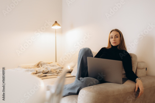 Concentrated millennial girl sit on couch working on laptop browsing internet at home during lazy weekend, focused young woman freelancer busy using computer surfing wireless web shopping online photo