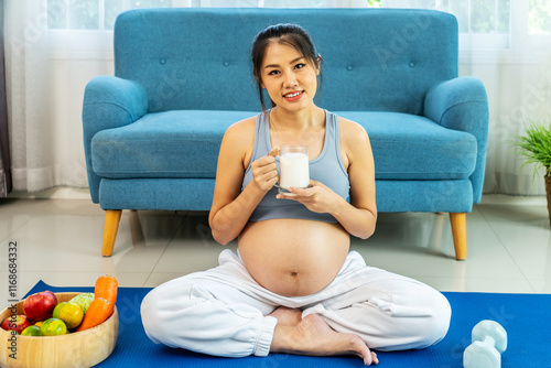 Eight months pregnant Asian woman wearing a sports bra sitting on a sofa stroking her belly and drinking calcium-fortified milk at living room at home, pregnancy and motherhood concept. photo