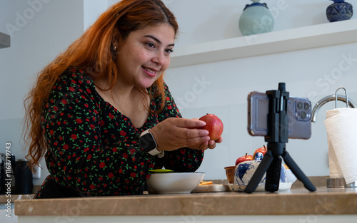 Close up of freckled young woman with red hair recording herself with her cell phone showing an apple making a dessert recipe at home photo