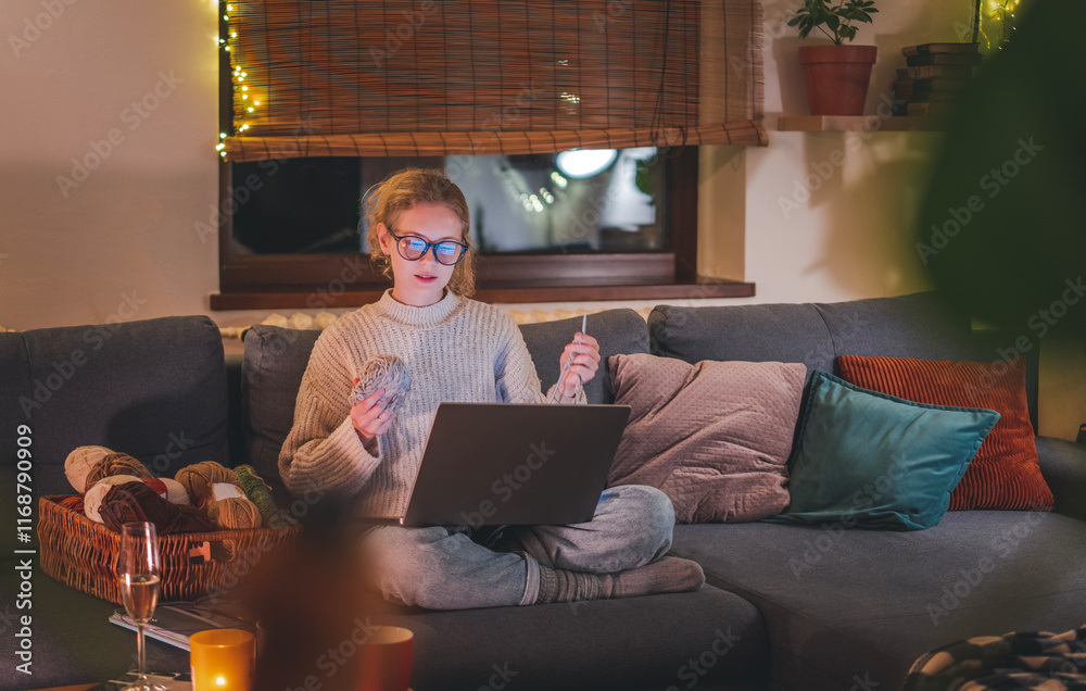 Young beautiful blonde woman with glasses learning to knit online sitting at home during Christmas holidays