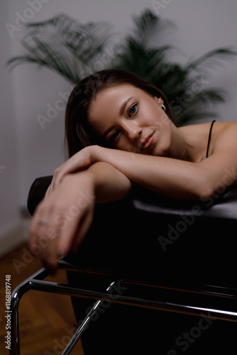 
This is an elegant portrait of a young woman with long, straight brown hair wearing a black dress. She is seated on a dark chair, slightly turned to the side, with her eyes closed and a subtle, seren