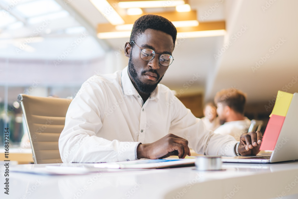 Fototapeta premium Concentrated african american male employee in eyewear for vision correction reading finance report and analyzing, serious dark skinned male manager sitting at working table with laptop computer