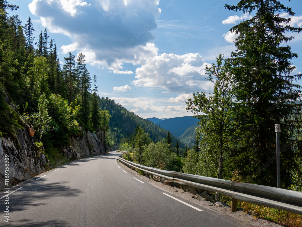 Fototapeta premium Scenic mountain road in Norway winding through lush greenery and under vibrant sky with scattered clouds.