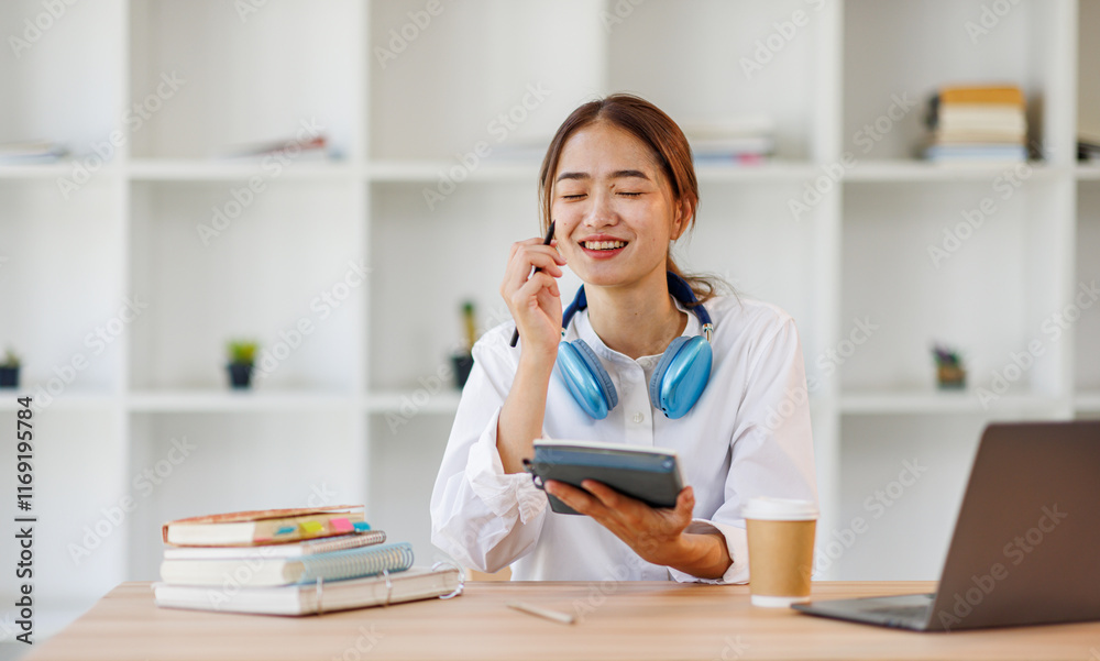 Concentrated young asian female student engaged in academic research with laptop and headphones at modern library desk, writing Notes and Studying Online.