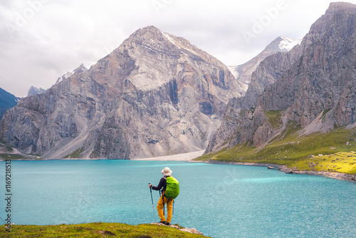 A woman stands on a rock with lush green grass by the Tientang Hu lake, Xinjiang, China. There are many mountains behind. The soft cool light shines on the still and shimmering blue water surface.