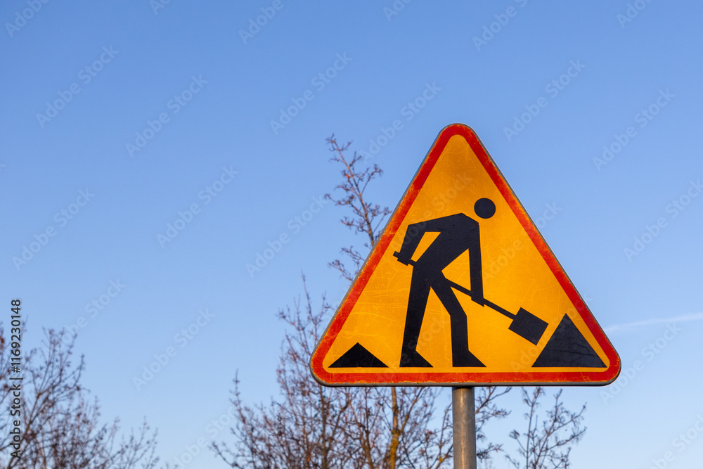 Road work warning sign against clear blue sky, triangular yellow traffic sign with worker symbol indicating construction zone ahead Road work warning sign against clear blue sky, triangular yellow traffic sign with worker symbol indicating construction zone ahead