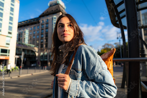 A curious woman carrying a denim jacket and a backpack walking through the busty city and looking aside.