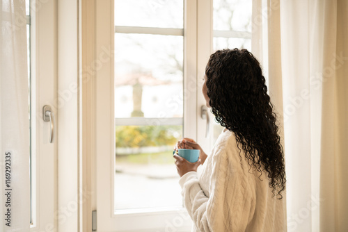 Woman relaxing in bathrobe, sipping coffee near window, soft morning light illuminating tranquil garden landscape