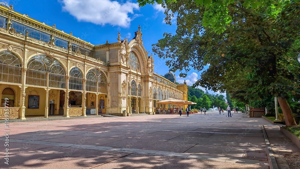 Fototapeta premium The Maxim Gorky Colonnade in Mariánske Lazne, Czech Republic with ornate architecture, large arched windows, and intricate details, surrounded by greenery and a bright blue sky.