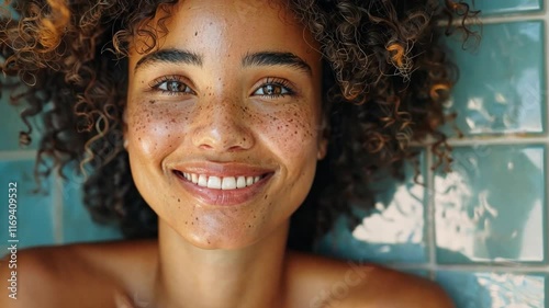 A young woman with curly hair and freckles smiles joyfully while resting her hand on her chin, surrounded by a calm atmosphere at home. Natural light enhances her radiant skin.