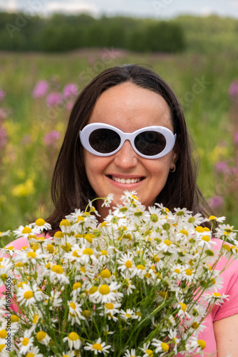 A joyful young woman wears white sunglasses while holding a fragrant bouquet of daisies, surrounded by a vibrant field of blooming flowers on a sunny day. 