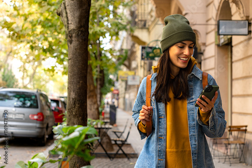 The woman walks through the city, checking her phone while carrying a backpack