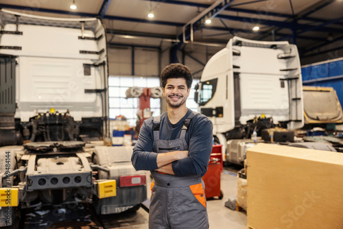 Professional confident multiracial serviceman standing with arms crossed in garage with trucks in background.
