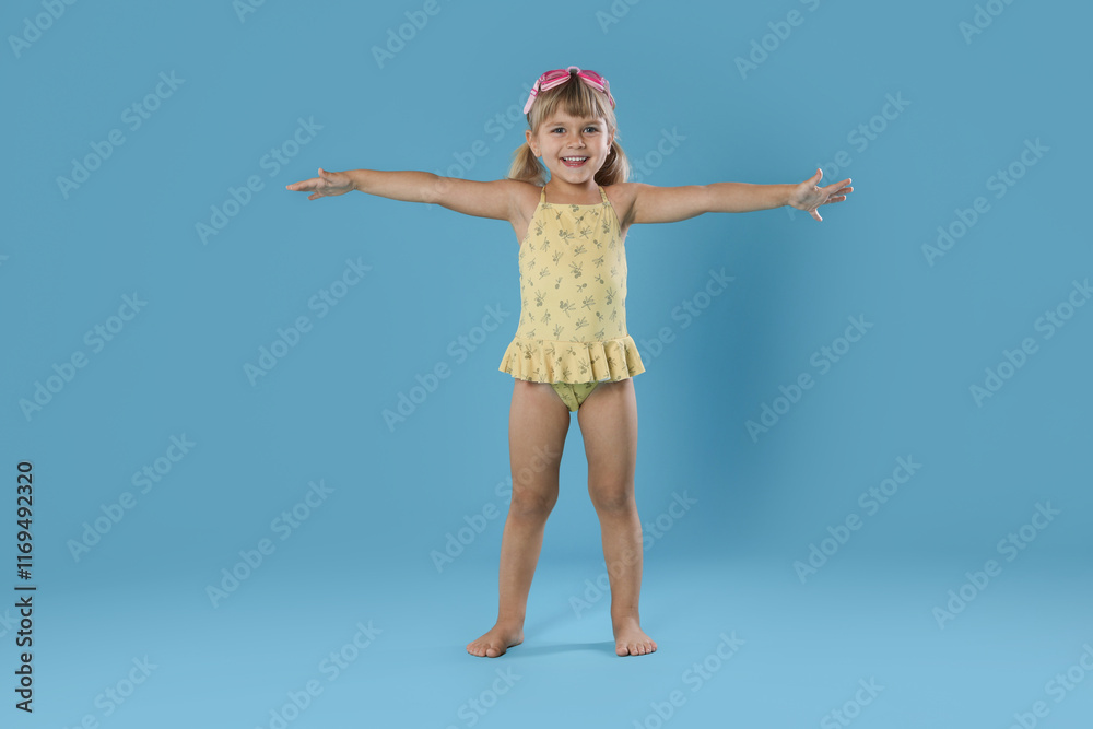 Cute little girl in swimsuit with swim goggles on light blue background