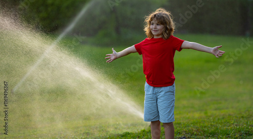 Cute little kid watering grass in the garden at summer day. Child play in summer backyard. Cute little boy is laughing and having fun running under water spraying sprinkler irrigation. Watering grass. photo