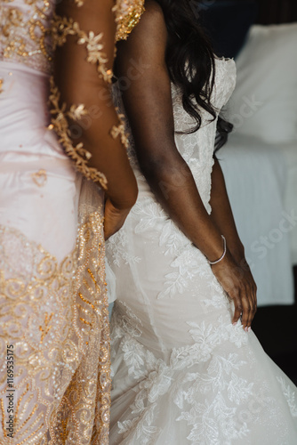 Close-up of a bride in a stunning lace wedding gown with floral details, assisted by a loved one in a gold-embellished outfit, showcasing intimate and elegant moments of wedding preparation