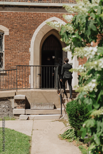 Historic brick church entrance with gothic arch and greenery in foreground, capturing the moment a man enters for a formal event under natural daylight