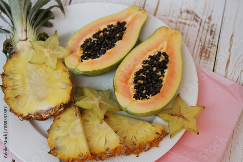 Fresh pineapple and papaya halves and carambola slices on the white plate on the wooden background photo