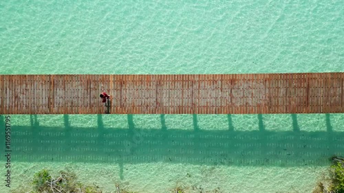 Toma aérea cenital de dron de joven y atractiva mujer en vestido rojo, caminando en un muelle de madera, en el agua color turquesa y los manglares del Parque Ecológico de Bacalar en México.