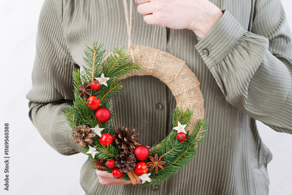 Woman holds a classic Christmas door wreath with fir twigs, red balls, small apples and cones.