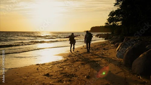 Hiking in sunset time, two tourists walking together on shore of beautiful river . Back view, slow motion with silhouette of travelers with backpacks strolling on sandy beach in evening, rear view